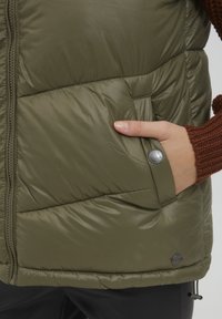 Gilet rembourré vert olive avec un motif matelassé, fermeture éclair et poche latérale dotée d'un bouton-pression. Le matériau semble brillant et lisse.