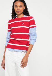 Red and white striped long-sleeve shirt with blue striped collar. Features logo on chest. Worn with white pants, focusing on layered design.