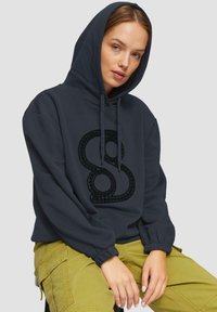 Hoodie noir foncé avec un grand logo en relief noir portant le chiffre 8. Il possède une capuche avec cordons de serrage et des poignets élastiques, associé à un pantalon cargo vert olive.