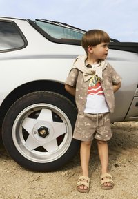 Enfant portant une chemise à manches courtes beige à motifs et un short, avec un t-shirt blanc à motifs, se tenant à côté d'une roue de voiture de luxe argentée.
