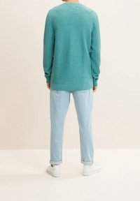 Personne debout de dos portant un pull texturé couleur sarcelle, un jean bleu clair retroussé et des baskets blanches sur un fond uni.