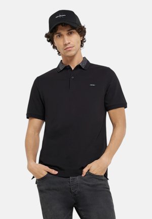 Uomo che indossa una polo nera con colletto a fantasia, jeans neri e un berretto da baseball nero, in piedi con le mani in tasca.