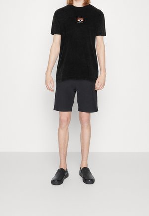 Camiseta negra de manga corta con un gráfico central que presenta un diseño de sol y ojo, combinada con pantalones cortos negros y zapatos negros sin cordones.