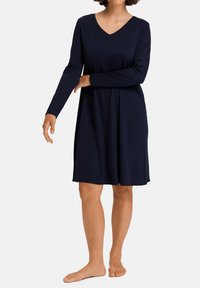 Donna in un vestito lungo fino al ginocchio a maniche lunghe di colore blu navy, in piedi a piedi nudi contro uno sfondo bianco semplice.