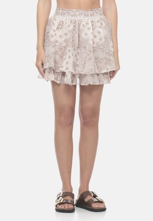 Donna che indossa una minigonna beige a strati con motivo paisley con fascia elastica in vita e sandali marroni con doppia fibbia, in piedi su sfondo bianco.
