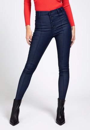 Femme portant un jean skinny bleu foncé taille haute avec boutons devant, associé à des bottines noires et un haut rouge côtelé à manches longues.