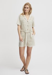 Combinaison en lin beige avec manches courtes, boutons sur le devant, col et ceinture à la taille. Dispose de deux poches et d'une coupe décontractée pour le confort.