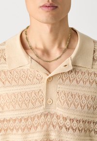 Beige strikket polo-skjorte med intrikat åpenvev, som har krage, to knapper og et delikat kjede.