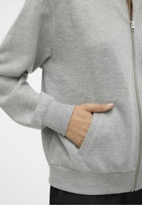 Szara bluza z kapturem zapinana na zamek, wykonana z miękkiego materiału, z prążkowanymi mankietami, kieszeniami po bokach i gładką fakturą. Projekt prosty i funkcjonalny.