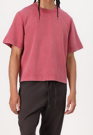 Person iført en løstsiddende falmet rød kortærmet t-shirt og sorte bukser med snor, vist fra nakke til midt på låret.