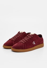 Bordowe zamszowe trampki z brązową gumową podeszwą, wyposażone w płaskie sznurowadła i biały logo po boku. Gładka tekstura i minimalistyczny design.