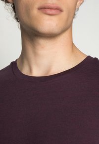 Gros plan sur le bas du visage et le cou d'une personne portant un t-shirt à col rond de couleur violet foncé, sur un fond uni clair.