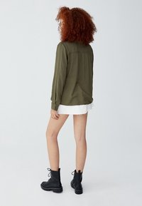Chemise vert olive à manches longues avec poignets à boutons, tissu texturé, coupe décontractée. Assortie à une jupe blanche effilochée et des bottines noires.