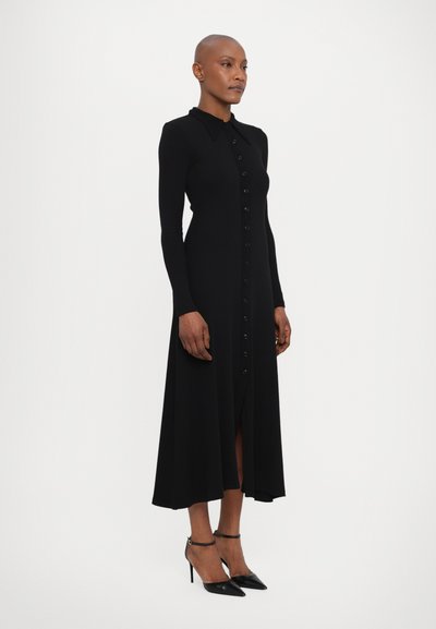 Melna, garām piedurknēm kleita ar pogu aizdari priekšā, apkakli un sānu šķēlumu, izgatavota no teksturēta auduma, kopā ar melniem augstiem papēžiem.