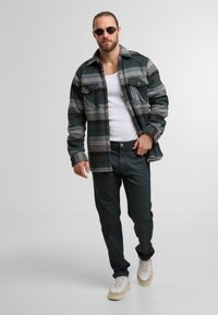 Uomo che indossa una giacca a quadri scuri con bottoni, una canotta bianca, pantaloni neri, sneaker bianche e occhiali da sole tondi che cammina in avanti.