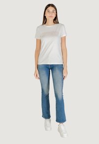 Camiseta blanca de manga corta con un logo "GUESS" texturizado, combinada con jeans acampanados de color azul claro y zapatillas blancas sobre un fondo neutro.