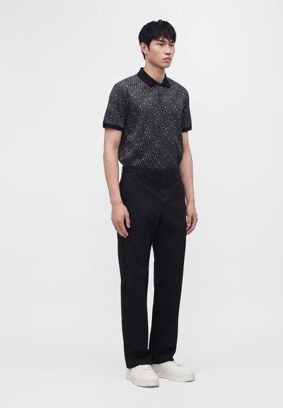 Jeune homme portant un polo à manches courtes noir et gris à motifs, un pantalon noir et des baskets blanches, debout devant un fond uni.