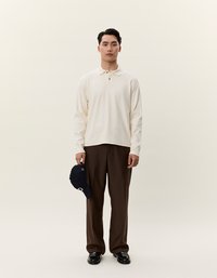 Világos krémszínű, hosszú ujjú pólóing, gallérral, sötétbarna nadrággal párosítva. A modell egy tengerészkék baseball sapkát tart. Fekete cipők egészítik ki a megjelenést.