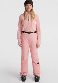 Růžový lyžařský outfit se zipovou pulovrem a širokými kalhotami s černým opaskem, doplněný černými lyžařskými botami a tmavou čelenkou.