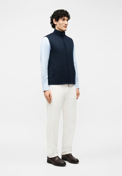 Άνδρας με σγουρά μαλλιά που φοράει ναυτικό μπλε γιλέκο με φερμουάρ πάνω από γαλάζιο ριγέ πουκάμισο, λευκό παντελόνι και σκούρα καφέ παπούτσια, στέκεται μπροστά σε ένα απλό φόντο.