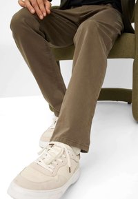 Bruine katoenen broek met een slim fit, gecombineerd met crèmekleurige sneakers met een gestructureerde bovenkant en een witte zool, zittend op een groene stoel.