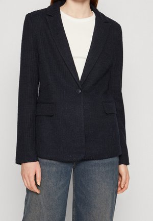 Blazer texturé bleu marine avec deux poches avant, fermeture à un bouton et coupe ajustée. Porté par-dessus un haut blanc, il présente un col cranté.
