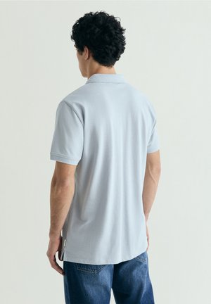 Hombre con cabello rizado y oscuro de espaldas, vestido con una camisa polo de manga corta azul claro y vaqueros azules sobre un fondo liso.