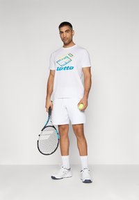 Hvid bomulds t-shirt med farverig grafisk design, hvide shorts, hvide sportssko med blå accenter, holder en tennisketsjer og bold.