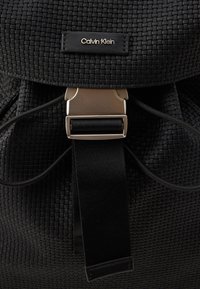 Juodas austo audinio kuprinė su logotipo etikete, metaline uždarymo detale ir tekstūruotu paviršiumi, reguliuojamais diržais.