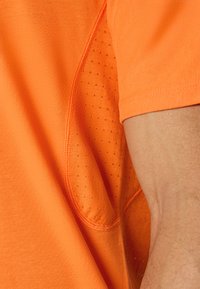 T-shirt a maniche corte in tessuto arancione brillante, con un dettaglio in pannello a rete sul lato, texture liscia e cuciture a contrasto.
