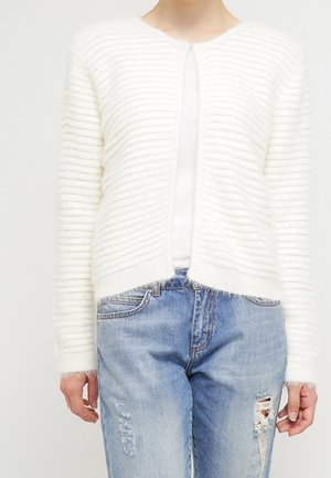 Personne portant un cardigan texturé blanc par-dessus un top blanc, assorti à un jean bleu clair déchiré, montré du cou jusqu’aux hanches.