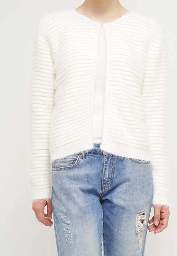 Personne portant un cardigan texturé blanc par-dessus un top blanc, assorti à un jean bleu clair déchiré, montré du cou jusqu’aux hanches.