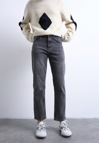 Άτομο που φοράει εκρού πουλόβερ με μαύρα διαμάντια, ψηλόμεσα σκούρα γκρι τζιν και λευκά αθλητικά παπούτσια, στέκεται σε γκρι δάπεδο.