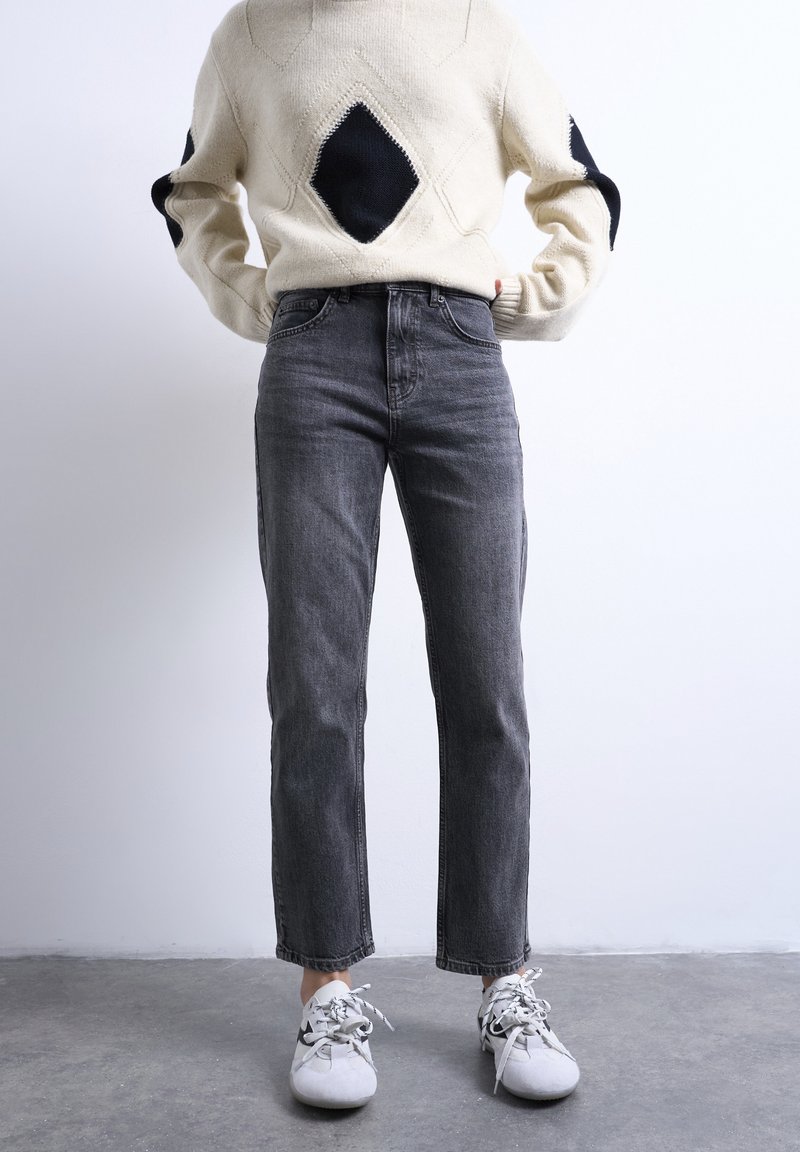 Άτομο που φοράει εκρού πουλόβερ με μαύρα διαμάντια, ψηλόμεσα σκούρα γκρι τζιν και λευκά αθλητικά παπούτσια, στέκεται σε γκρι δάπεδο.