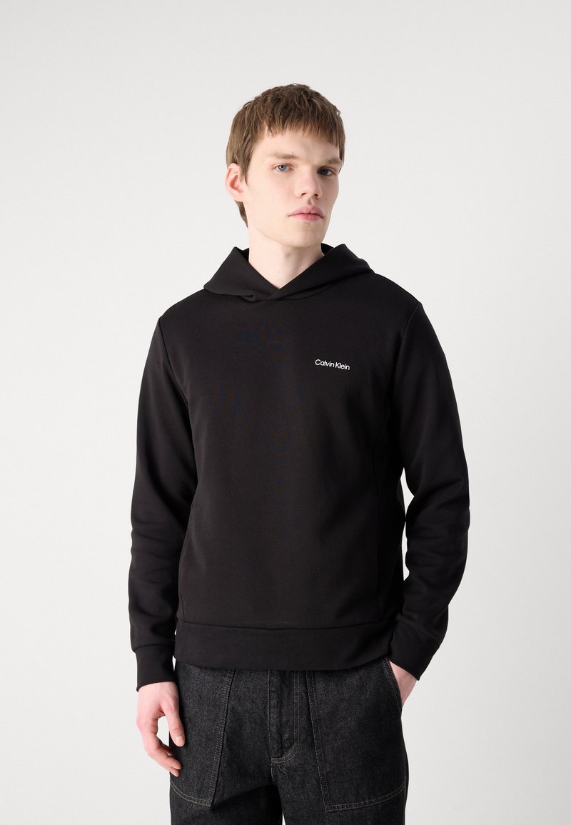 Jonge man die een zwarte Calvin Klein hoodie en zwarte jeans draagt, staande met één hand in zijn zak tegen een effen witte achtergrond.