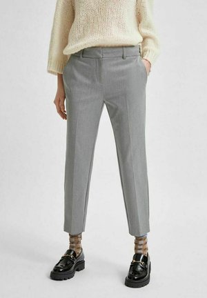 Henkilö pukeutuneena vaaleanharmaisiin räätälöityihin housuihin, beigenruudullisiin sukkiin, mustiin loafer-kenkiin ja kermaiseen neulepaitaan, kädet taskuissa.