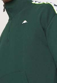 Pull en vert foncé avec un col montant, devant zippé sur moitié, logo blanc, et accents d'épaule blancs à motifs avec une finition néon. Tissu doux au toucher.