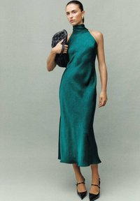 Vestido de satén verde azulado con cuello alto y una abertura lateral, con una textura sutil. Combinado con un bolso de mano negro trenzado y tacones apuntados negros.