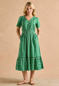Zelené potištené šaty s krátkymi rukávmi, priliehavým pásom a rozšírenou sukňou. Obsahujú tiered lem a gombíkovú výzdobu na prednej strane.