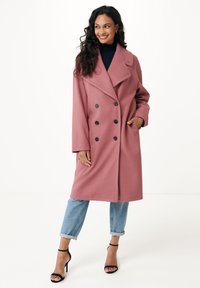 Cappotto in lana rosa chiaro con design doppiopetto, ampi revers, bottoni neri e tasche laterali, abbinato a jeans blu arrotolati e tacchi neri.