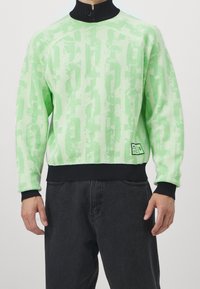 Uomo che indossa un maglione a motivi verde chiaro con colletto, polsini e orlo neri, abbinato a pantaloni neri larghi.