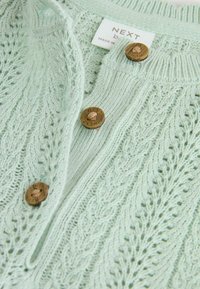 Gaiši piparmētru zaļš adīts kardigans ar kabeli rakstu, trīs brūniem pogām un struktūru, lai pievienotu detaļas.