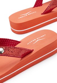 Red flip-flops with a textured strap, featuring a shiny accent and orange platform with white stripes. Brand name "Venice Beach" printed.