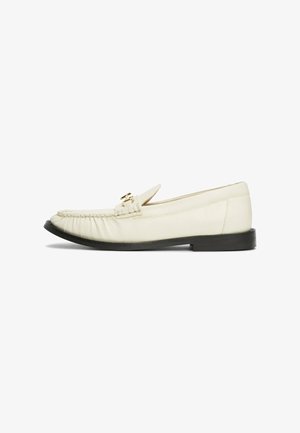 Mocassins en cuir crème avec un bout arrondi, présentant une boucle décorative en ton doré et un détail froncé à l'avant, reposant sur une semelle noire.