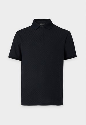 Homme portant un polo noir à manches courtes, un jean bleu clair à jambes larges avec les mains dans les poches et des baskets noires, debout devant un fond uni.