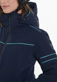Mörkblå jacka med hög krage, turkosa detaljer och dragkedja framtill, tillverkad i ett slätt yttertyg med matt finish.