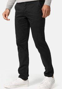 Black cotton trousers with a slim fit, flat front, and pockets, paired with white sneakers. Fabric has a smooth texture.