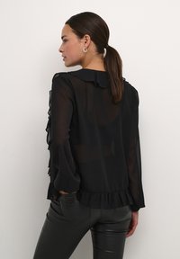 Sort gennemsigtig bluse med flæsekrave og manchetter, lange ærmer og en samlet kant; bæres med sorte tætsiddende bukser. Simpelt og elegant design.