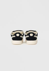 Juodos zomšos sportbačiai su auksiniu logotipu ir tekstūruota grietinėlės spalvos padu. Yra raštuota šoninė panelė ir minkšta vidaus apdaila, užtikrinanti komfortą.