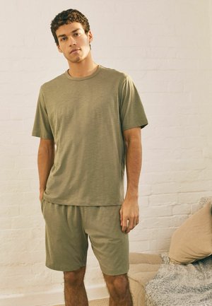 Nuori mies oliivinvihreässä lyhythihaisessa T-paidassa ja siihen sopivissa shortseissa seisoo sisätiloissa valkoista tiiliseinää vasten, sohvalla lähistöllä.